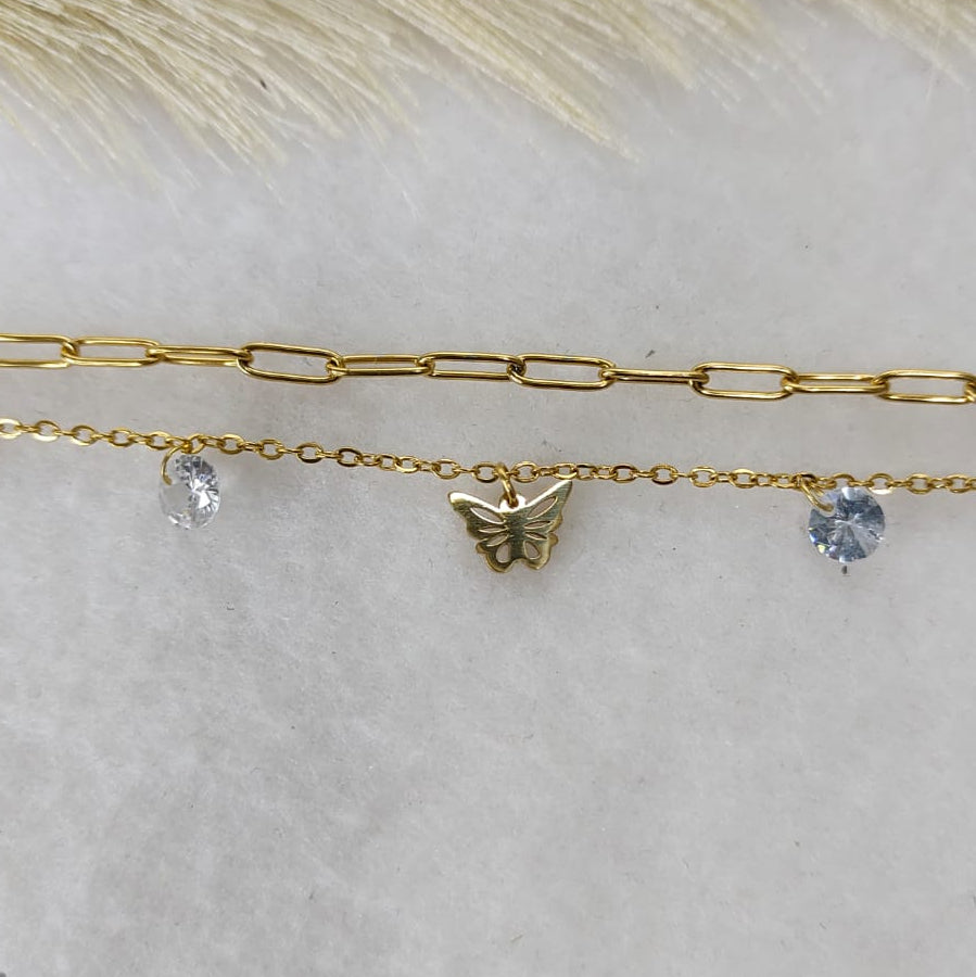 Two gold-toned chain necklaces laid flat on a light neutral surface, one with a paperclip link pattern and one with a delicate chain, a small butterfly charm, and clear stones.